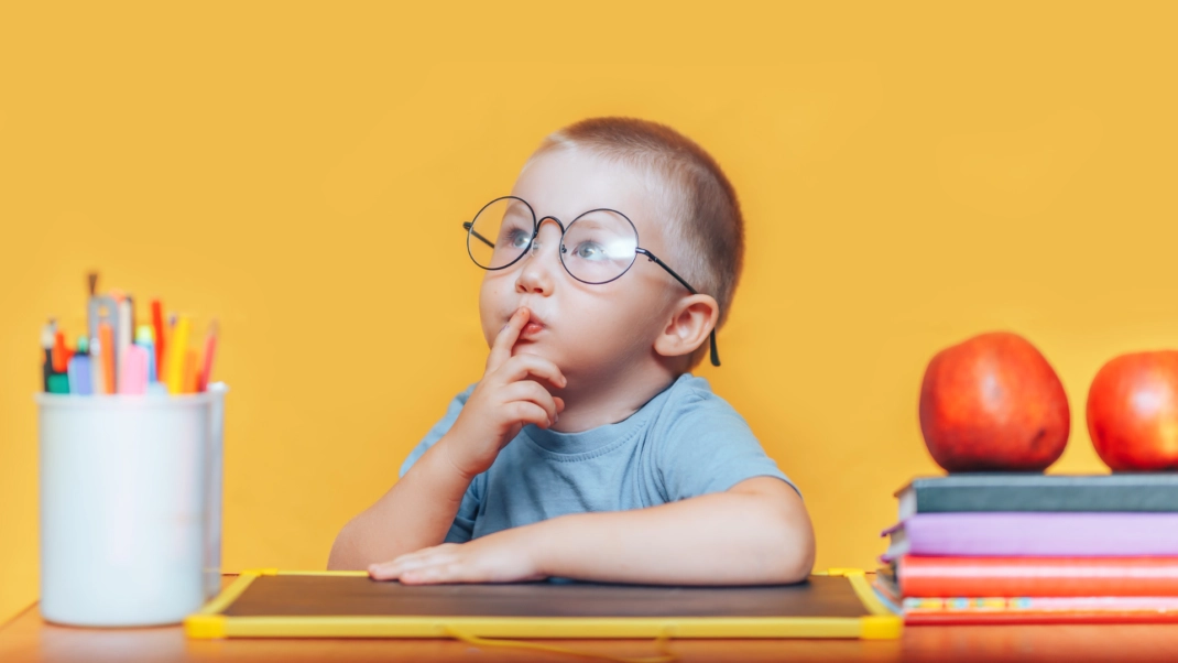 A young child wearing glasses sits at a desk with a finger on their lips, surrounded by colored pencils, apples, and stacked books, against an orange background.