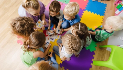 A group of young children sit on colorful foam mats, gathered around an adult, holding letter cards and engaging in a learning activity.