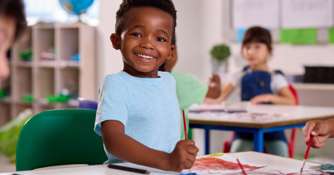 Child in a blue shirt smiling and painting a picture.