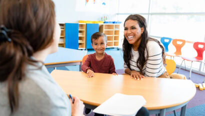 The cute preschool age boy watches quietly as his mid adult mom and his unrecognizable female teacher smile and laugh during the parent teacher conference.