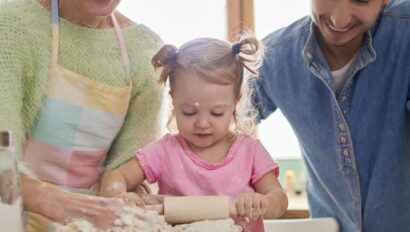A young child uses a rolling pin on dough with the help of two adults, likely her parents, in a bright kitchen setting.