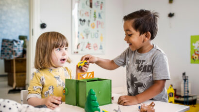 Two young children play with toy cars and animal figures at a table in a brightly decorated room with alphabet art on the wall.