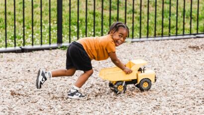 Child in an orange shirt pushing a toy truck outside.